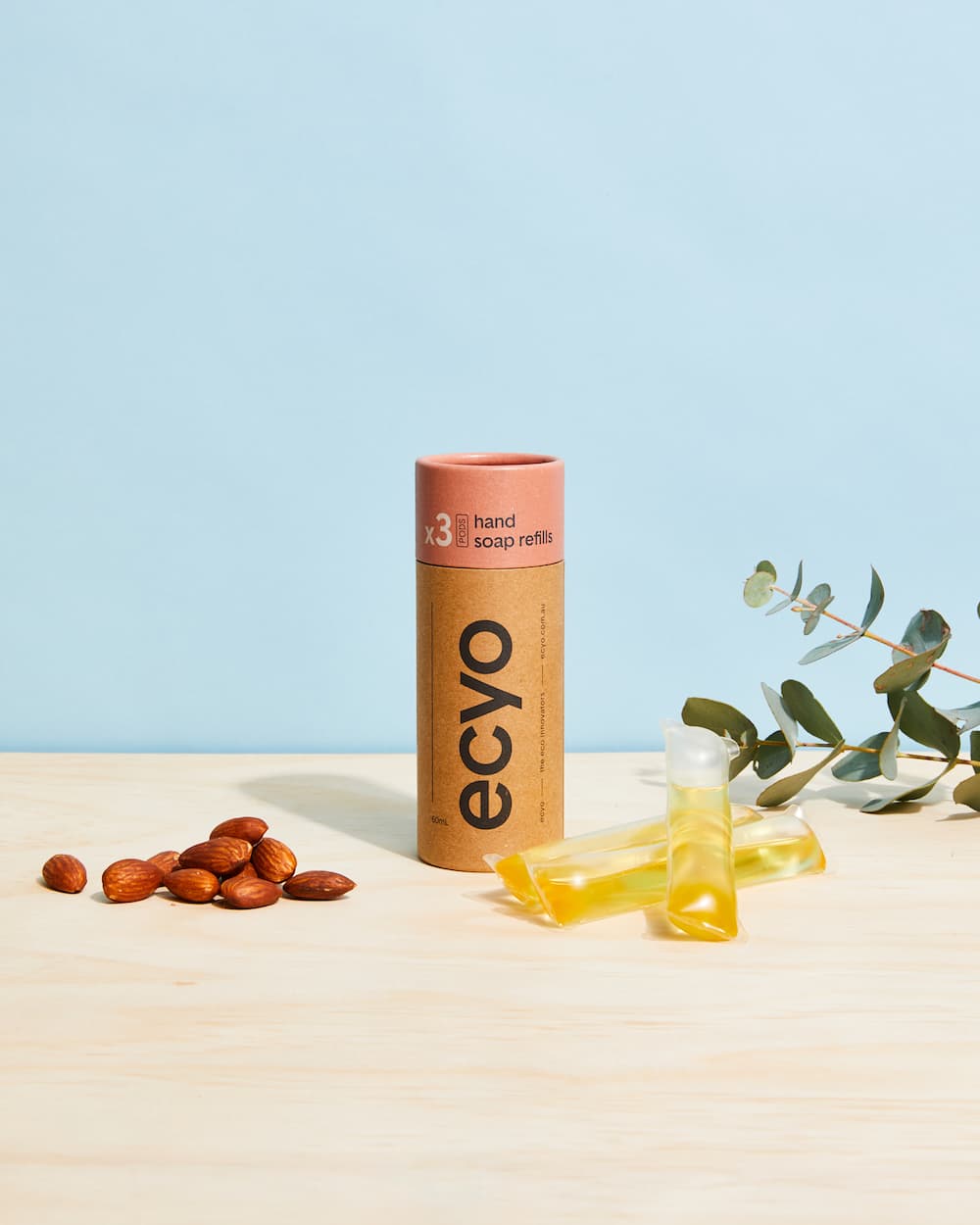 Ecyo hand soap refills on a light surface with almonds and eucalyptus leaves against a blue background