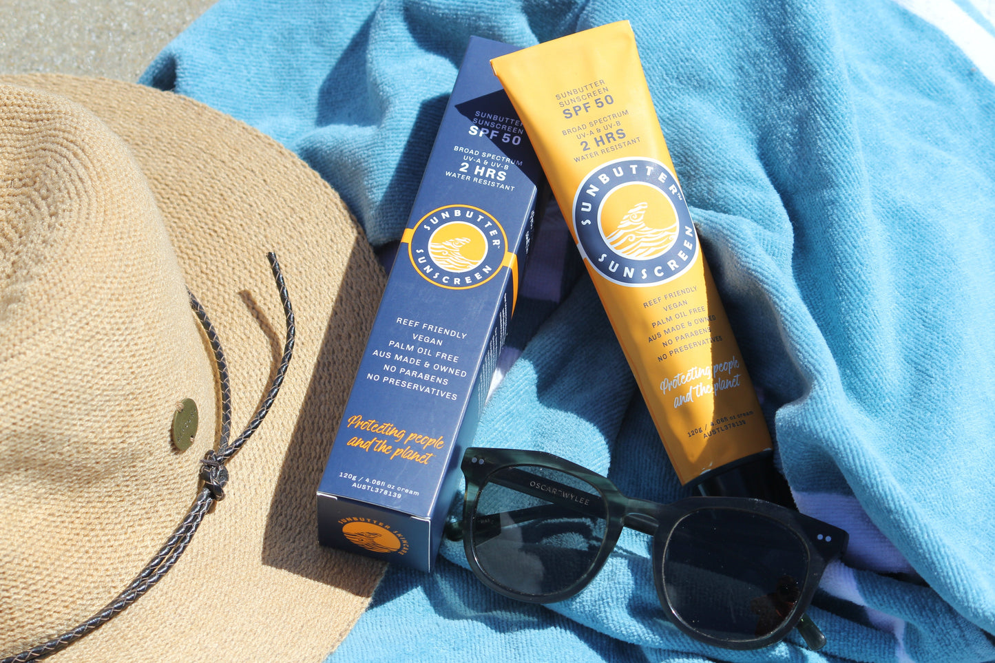 Sunbutter sunscreen tube, bottle, sunglasses, and hat on a blue towel