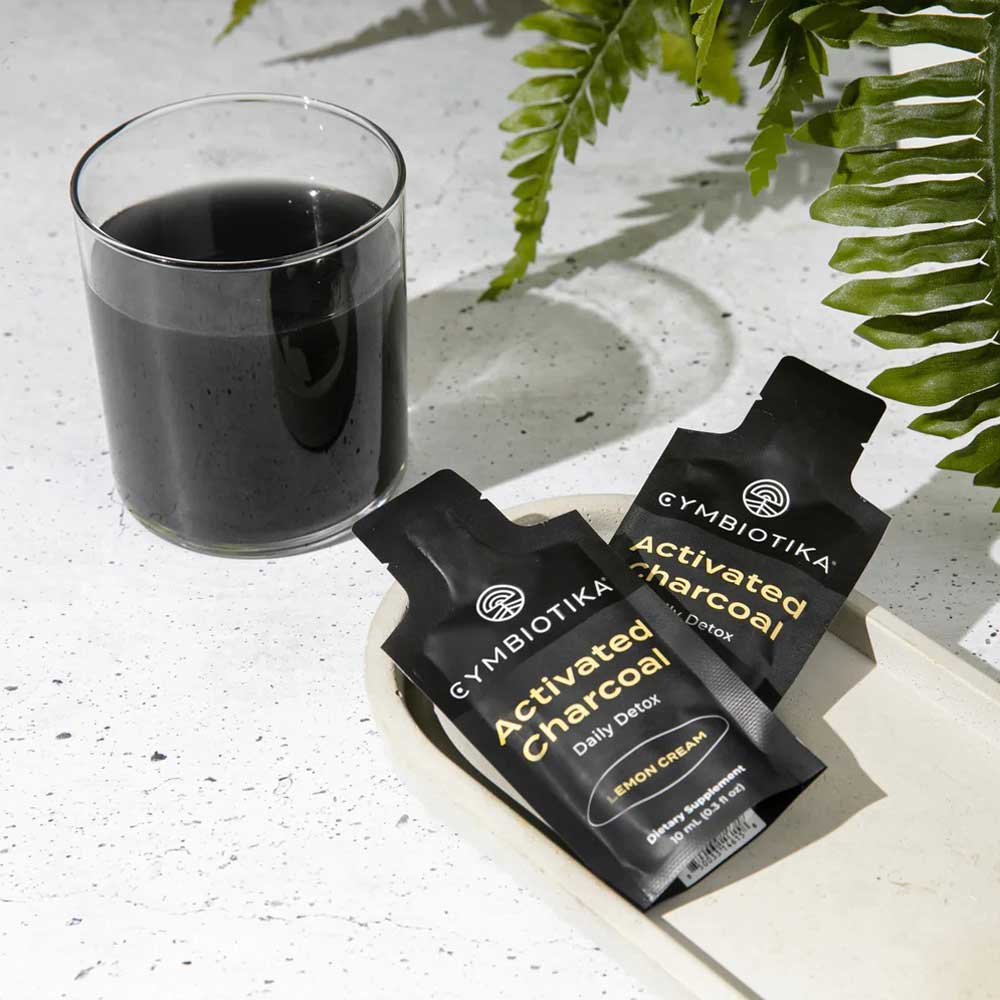 Gymbiotika Activated Charcoal packets with a glass of black liquid on a light surface with fern leaves.