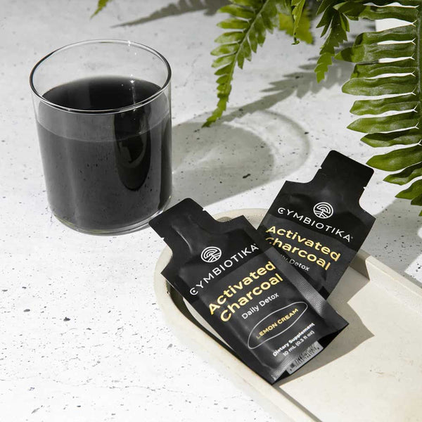 Gymbiotika Activated Charcoal packets with a glass of black liquid on a light surface with fern leaves.