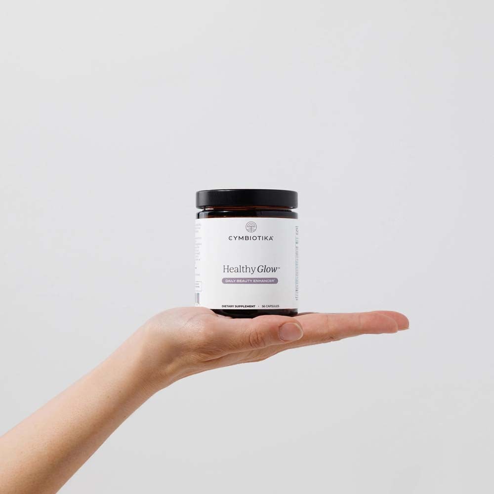 Hand holding a Cymbiotika Healthy Glow supplement jar against a light gray background