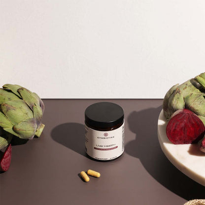 Supplement jar labeled 'Cymbiotika' with two capsules on a surface with artichokes and beetroot.