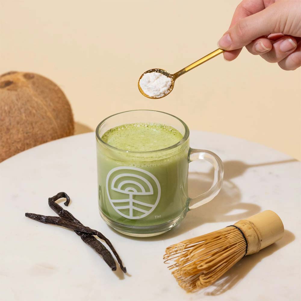 Green matcha drink with a spoon of nootropic creamer, vanilla beans, and a whisk on a light surface.