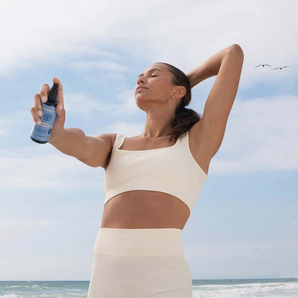 Woman applying magnesium oil spray on a beach with a clear sky