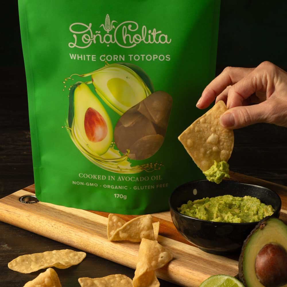 Green bag of Dona Cholita white corn totopos with a hand dipping a tortilla chip into guacamole.
