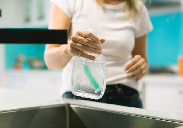 Person holding a bottle of cleaning solution with a green brush inside, in a bathroom setting.