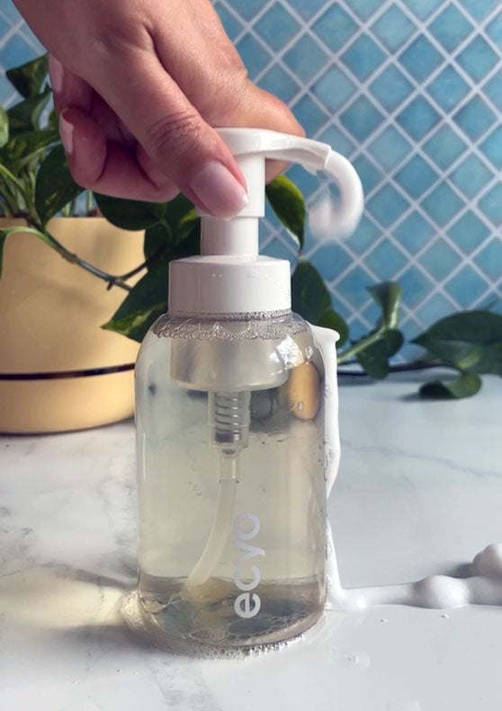 Hand holding a clear bottle with a pump, labeled 'ecyo', against a blue tiled wall.