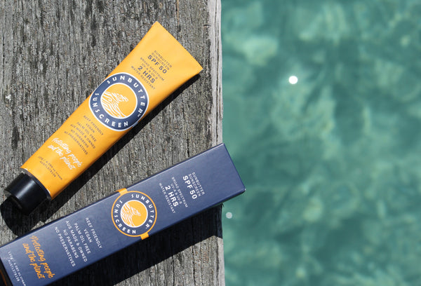Two tubes of sunbutter sunscreen, one yellow and one blue, leaning against a wooden post with water in the background.