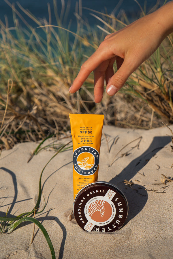  Sunbutter sunscreen tube and container on sand with grass and water in the background