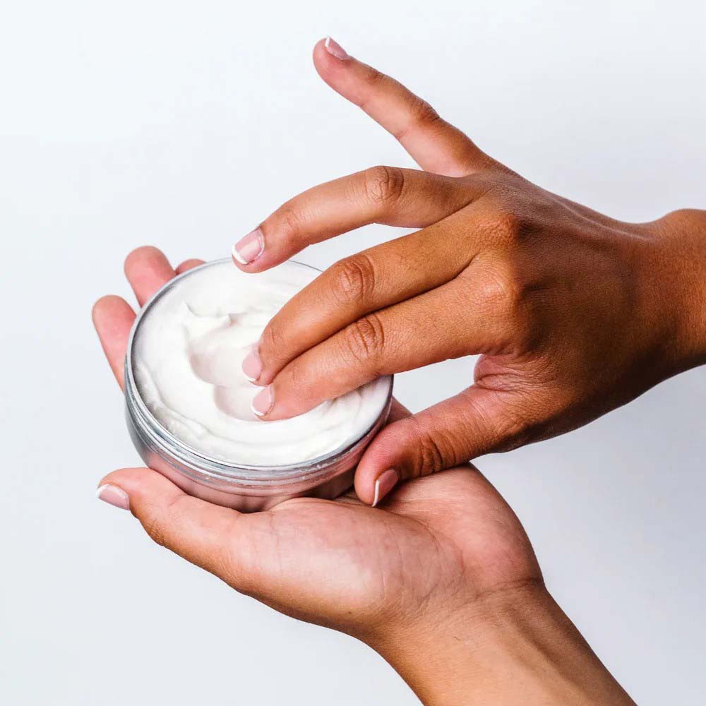Hand applying white cream from a jar against a light background
