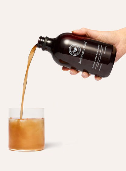 Hand pouring a brown liquid from a bottle labeled the beauty chef antioxidant boost into a glass on a white background.