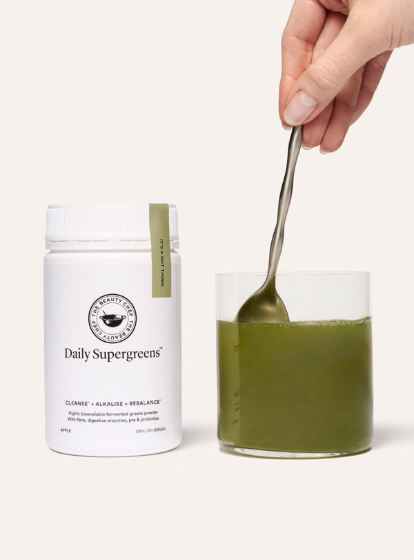 Container of 'Daily Supergreens' with a glass of green liquid and spoon on a white background