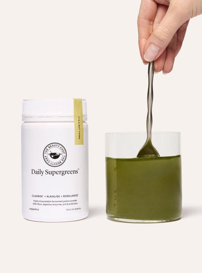 Daily Supergreens powder with a glass of green liquid and a spoon on a white background