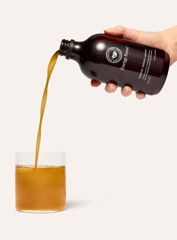 Hand pouring a brown liquid from a bottle labeled 'Energy Boost' into a glass on a white background.