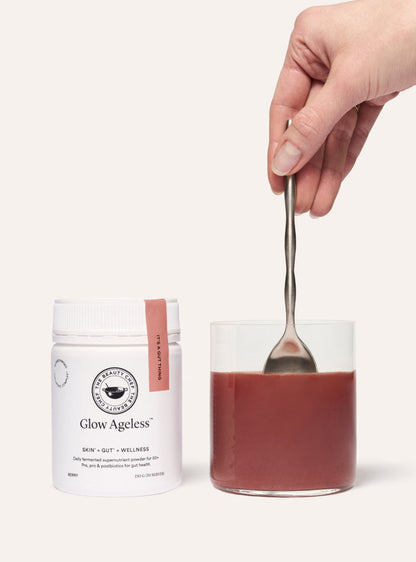 Hand holding a spoon over a jar of red powder with 'Glow Ageless' packaging on a white background