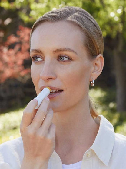 Woman outdoors applying Lavera Basis Sensitiv Lip Balm directly to her lips while wearing a white shirt and silver hoop earrings.