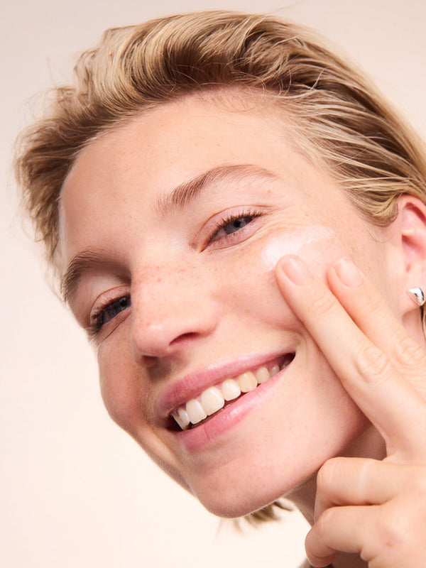 Smiling woman applying Lavera Basis Sensitiv Moisturising Cream to her cheek with her fingertips.
