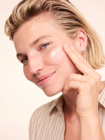 Woman smiling while applying a white streak of Lavera Basis Sensitiv Q10 Moisturising Day Cream to her cheek with one fingertip.