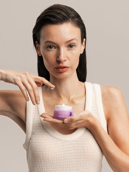 Woman holding a jar of Lavera Firming Day Cream while touching the cream with one fingertip.