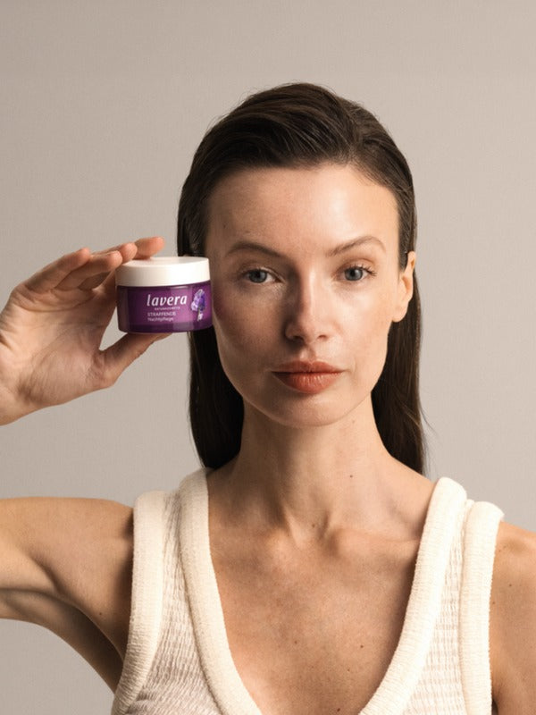 Woman holding a purple jar of Lavera Firming Night Cream beside her face against a warm neutral background.